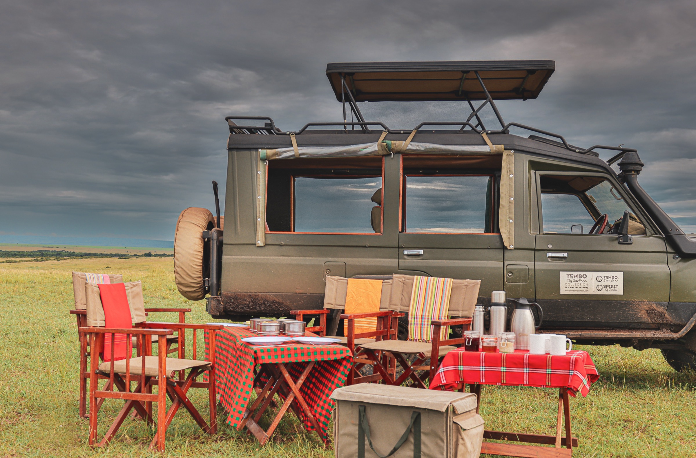 Bush breakfast set out on the grass beside an open-roofed safari vehicle under grey clouds: wooden director's chairs, a table laid with plates and steel food tins, and a second table with flasks, jars and white mugs