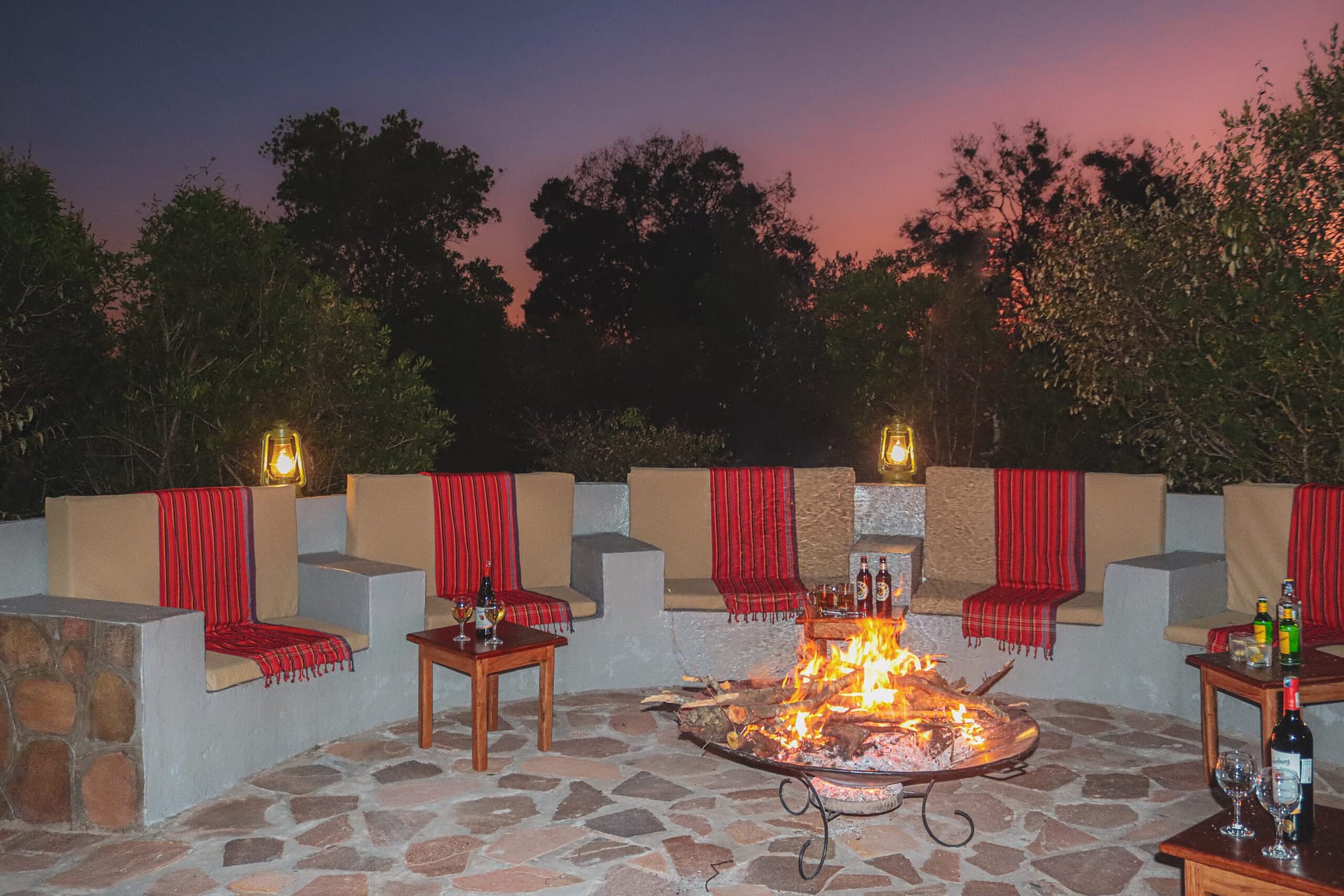 Circular stone fire pit lounge at dusk, with a log fire, two lit lanterns and red checked blankets over the cushioned seats