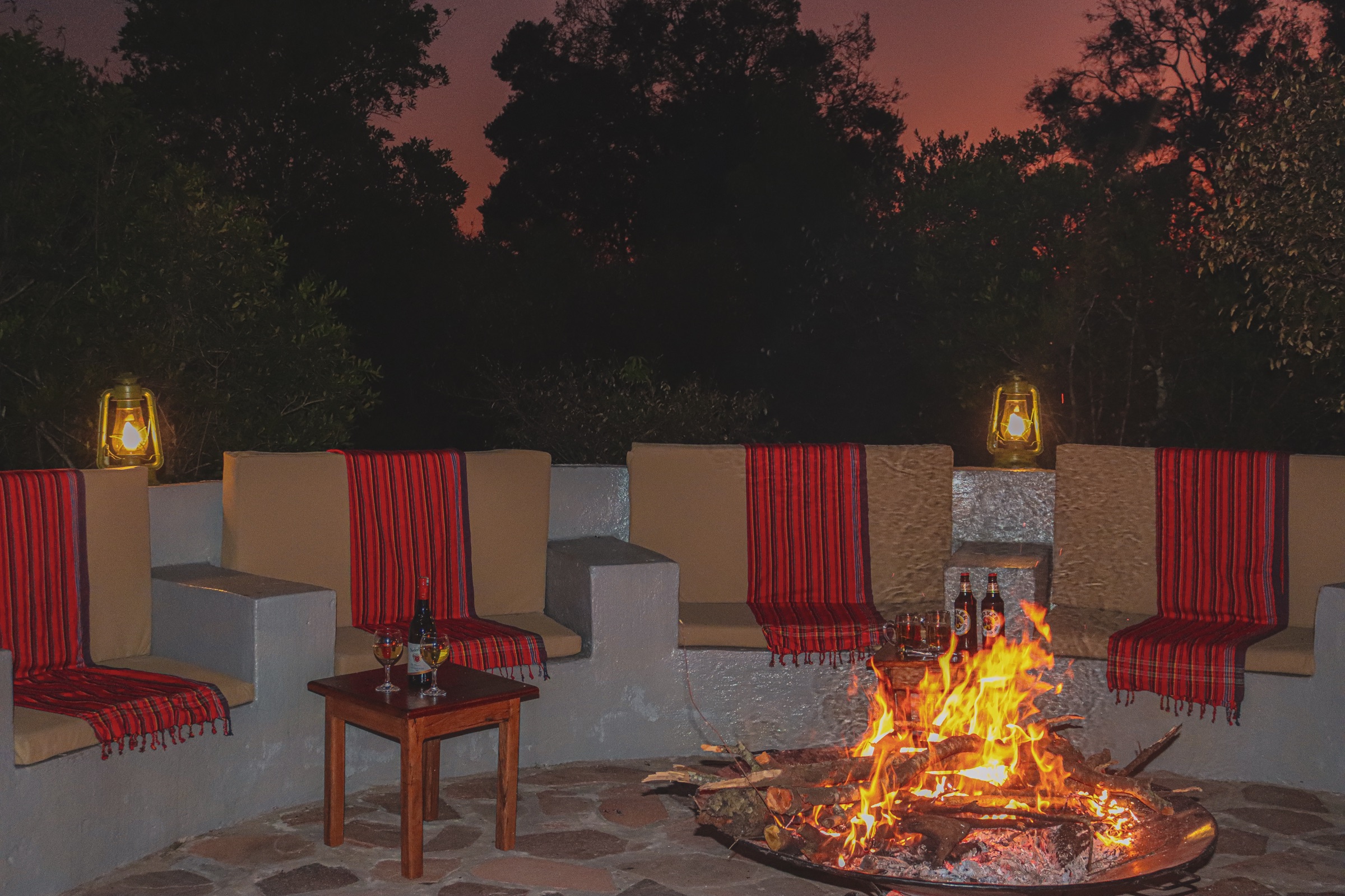 Log fire burning in a metal bowl beside curved built-in seats at dusk, with red striped blankets on the cushions, two lit lanterns, white wine on a small wooden table and two beers by the fire