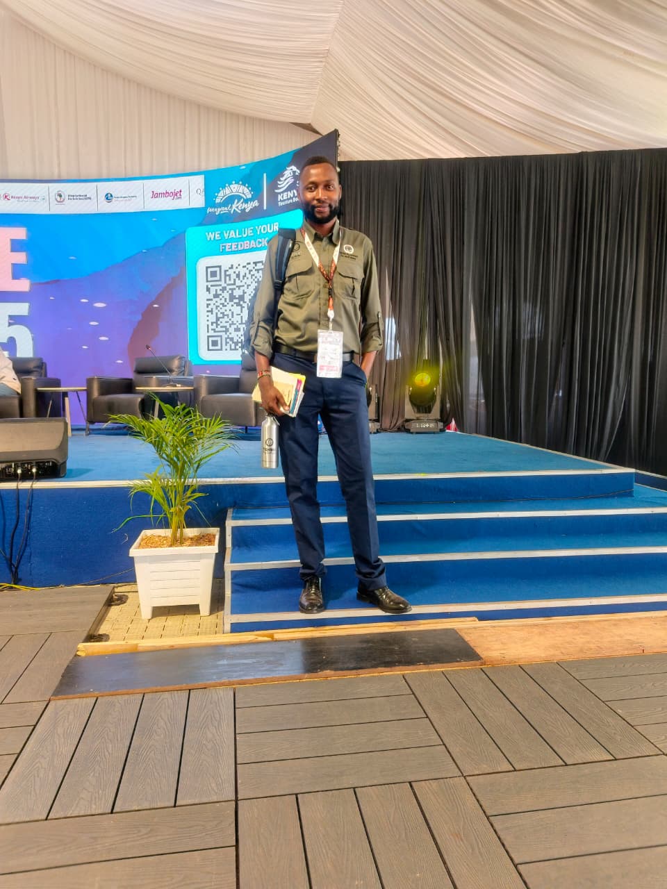 A man in an olive shirt and navy trousers, holding papers and a steel water bottle, standing on the blue steps of a conference stage inside a marquee, with a Magical Kenya screen behind him