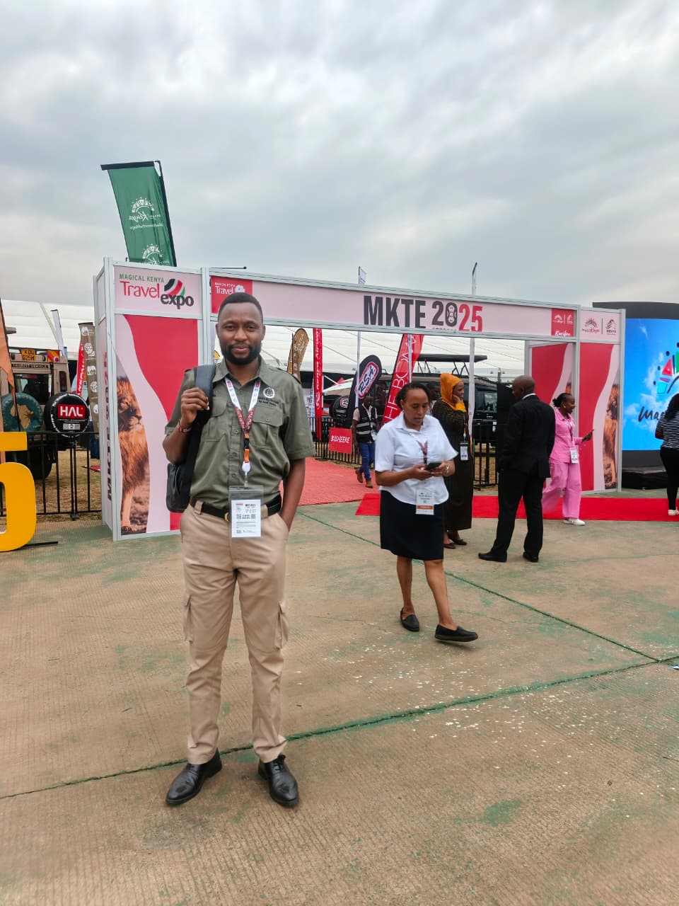 A man in an olive shirt and khaki trousers, with an expo badge and a backpack, standing in front of the MKTE 2025 entrance arch of the Magical Kenya Travel Expo as other visitors walk past