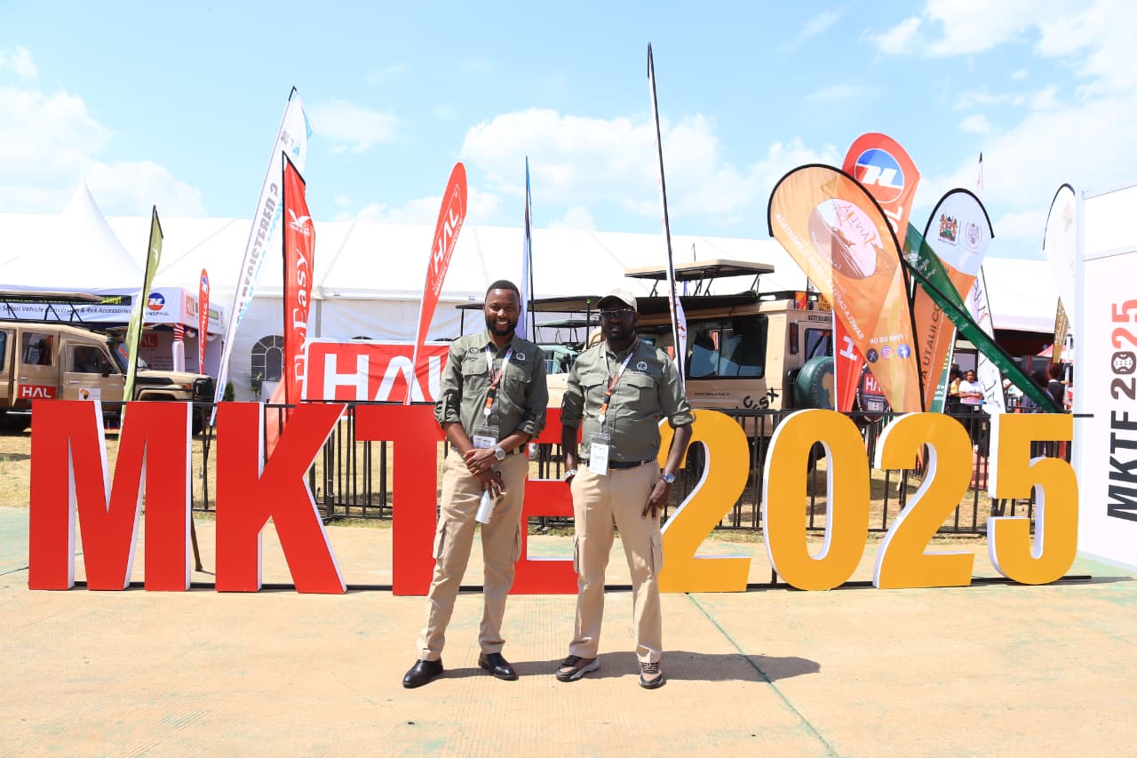 Two men in olive shirts and khaki trousers, wearing expo badges, standing in front of large red and yellow MKTE 2025 letters, with exhibitor flags and safari vehicles behind them