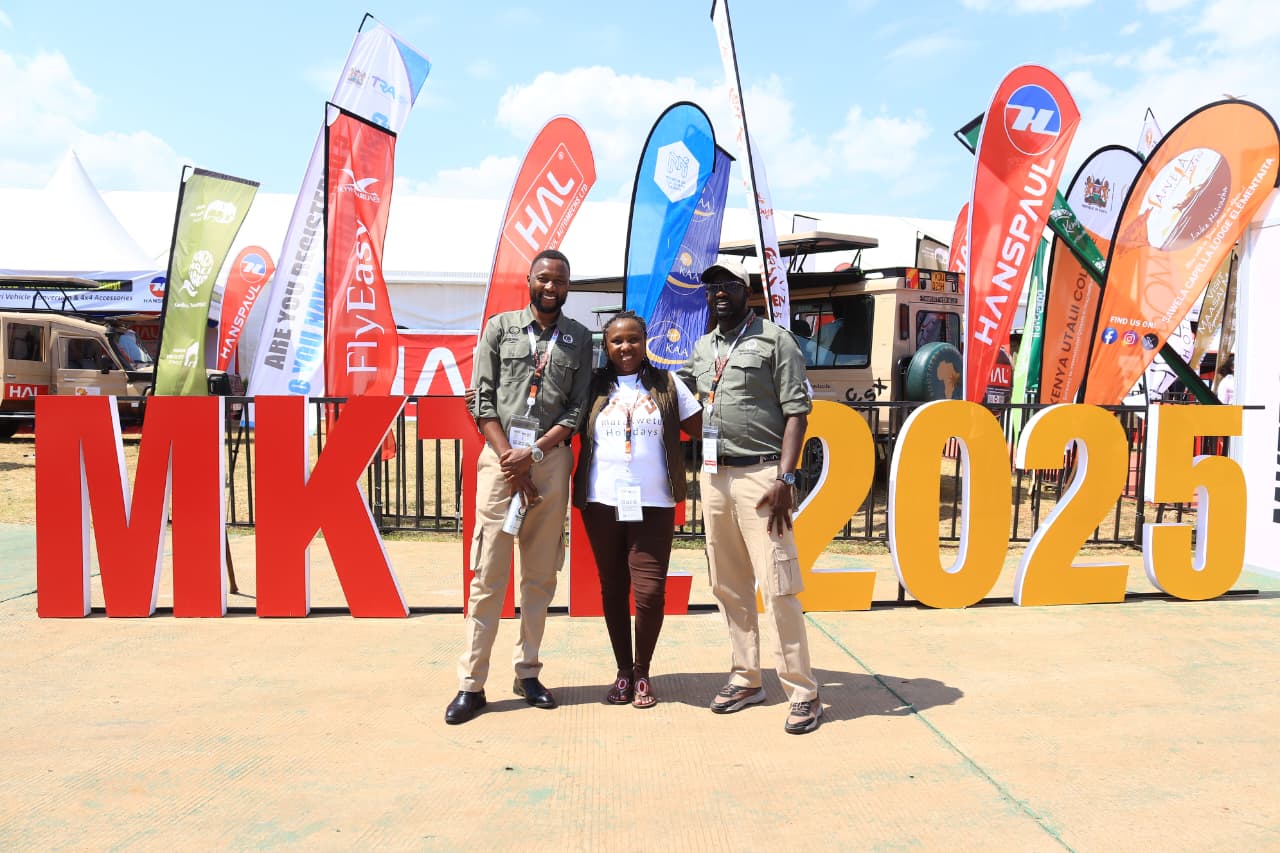 Two men in olive shirts and khaki trousers with a woman in a white T-shirt between them, standing in front of large red and yellow MKTE 2025 letters on a sunny day