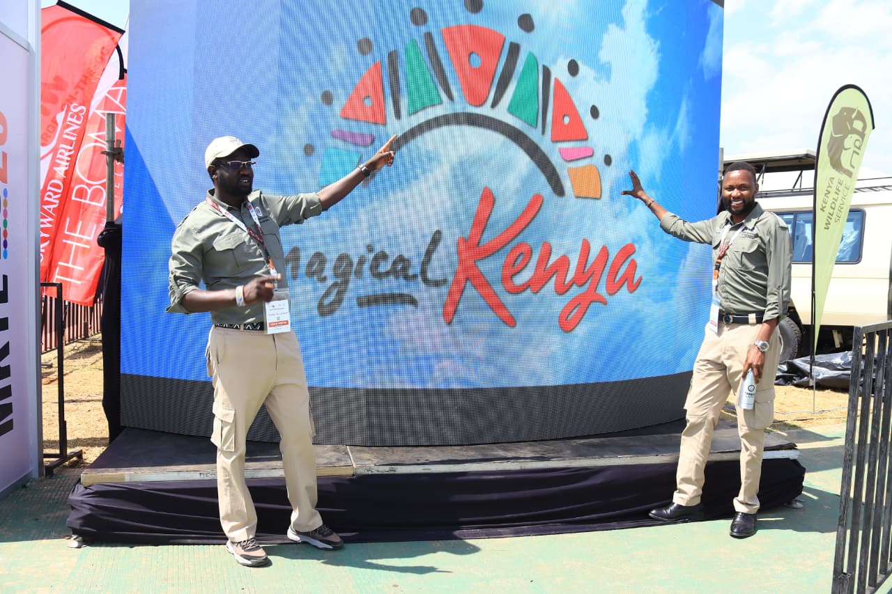 Two men in olive shirts, khaki trousers and expo badges pointing at the Magical Kenya logo on a large curved LED screen at the expo grounds