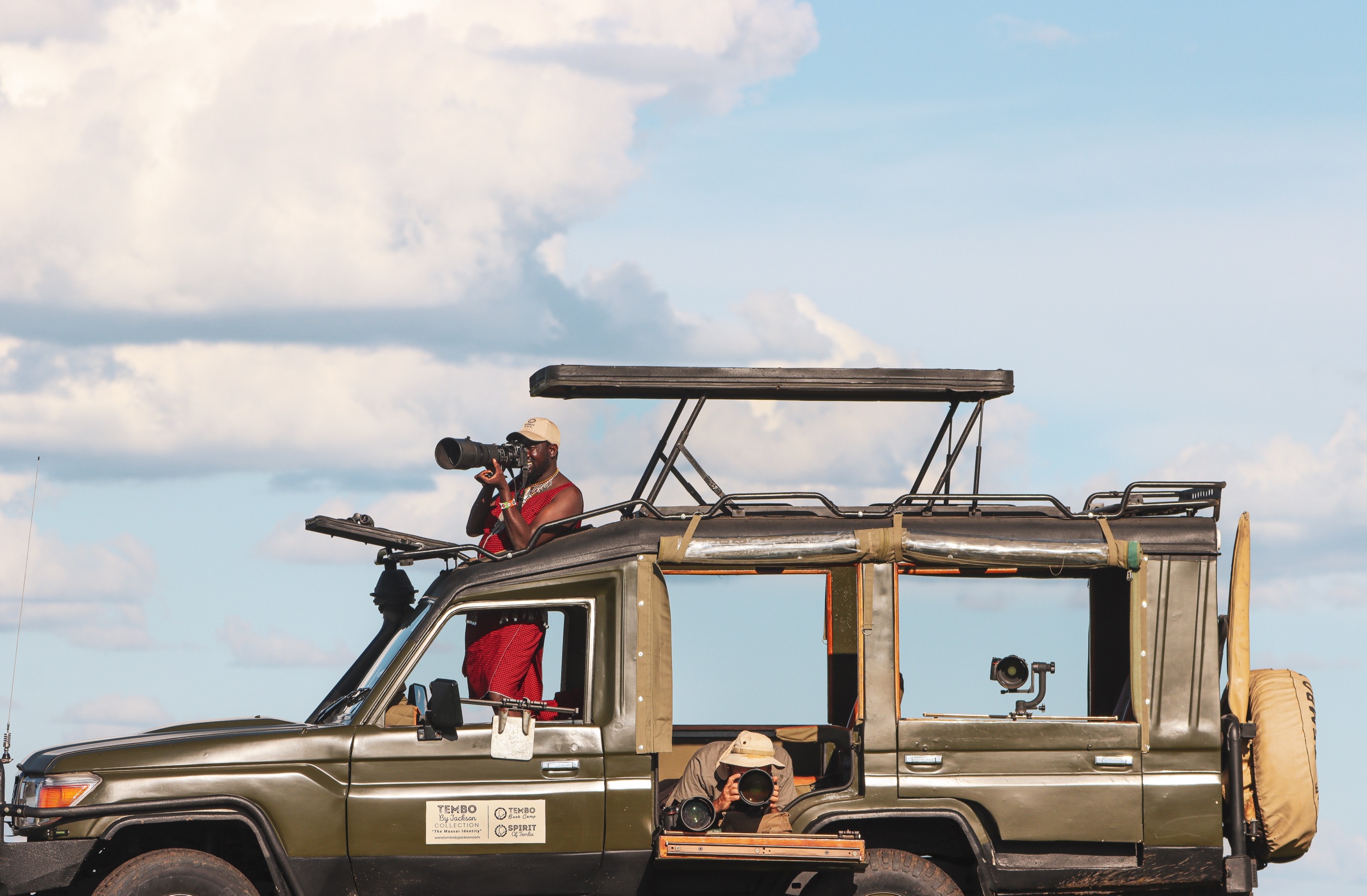 Safari vehicle with a raised roof under a cloudy blue sky: a host in a red shuka photographs with a long lens from the roof hatch, a guest in a bush hat shoots from a low side opening, and another long lens sits on a mount at the rear window
