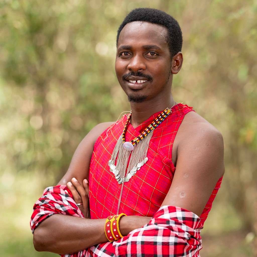 Smiling member of the Tembo team with folded arms, wearing a red checked shuka, a beaded collar with silver chains and beaded bracelets, in front of blurred green bush