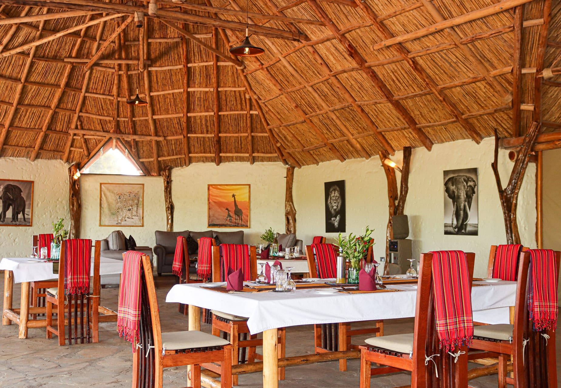 Dining room under a high thatched roof, with tables laid in white cloths, red checked blankets on the chairs and wildlife prints on the walls